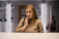 A woman in a brown sweater experiencing a toothache, holding her cheek indoors.